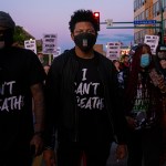 A black man standing in the center of a colorful photo. He wore a mask and a T-shirt, 