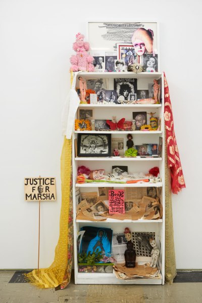 An altar-like sculpture in a white bookcase featuring various dolls, photographs, beads, and more. 