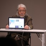 An elderly Asian man holding a laptop screen to the audience.