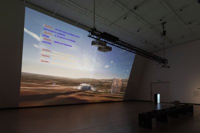 The view from the large screen in the museum gallery shows the landscape of the Golan Heights with chat text from video games. 